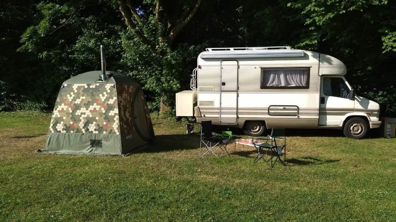 Mobiba MB-10 Saunazelt neben Wohnwagen Mobiba MB-10 Zeltsauna in Wabentarnung neben einem Wohnwagen auf einer Wiese; davor Campingstühle und Tisch.