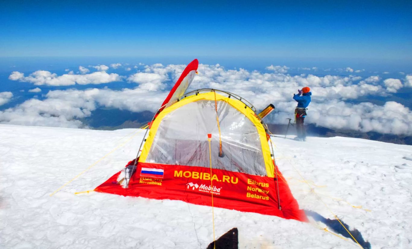 Mobiba Saunazelt am Elbrus (5640 m) Rot-gelbes Mobiba Saunazelt mit transparenten Fenstern auf schneebedecktem Gipfelplateau am Elbrus (5640 m); im Hintergrund Wolkenmeer.