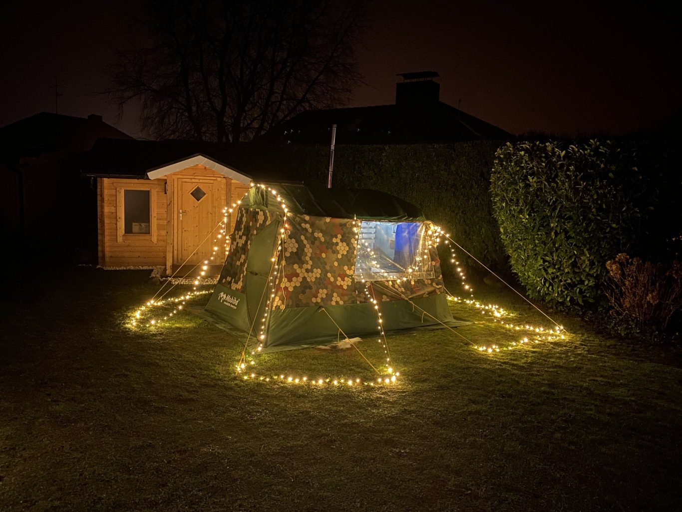 Mobiba MB-10A1 mit Überzelt ZP-10A1 im Garten Mobiba MB-10A1 Saunazelt mit Überzelt (Zusatzplane) ZP-10A1 in einem Garten bei Nacht, dekoriert mit Lichterkette.