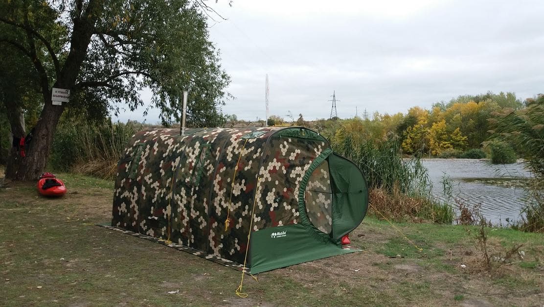 Saunazelt Mobiba MB-104 am Fluss. Mobiba MB-104 Saunazelt in Farbe Wabetarnung am Flussufer auf einer Wiese, im Hintergrund Bäume und Wasser.