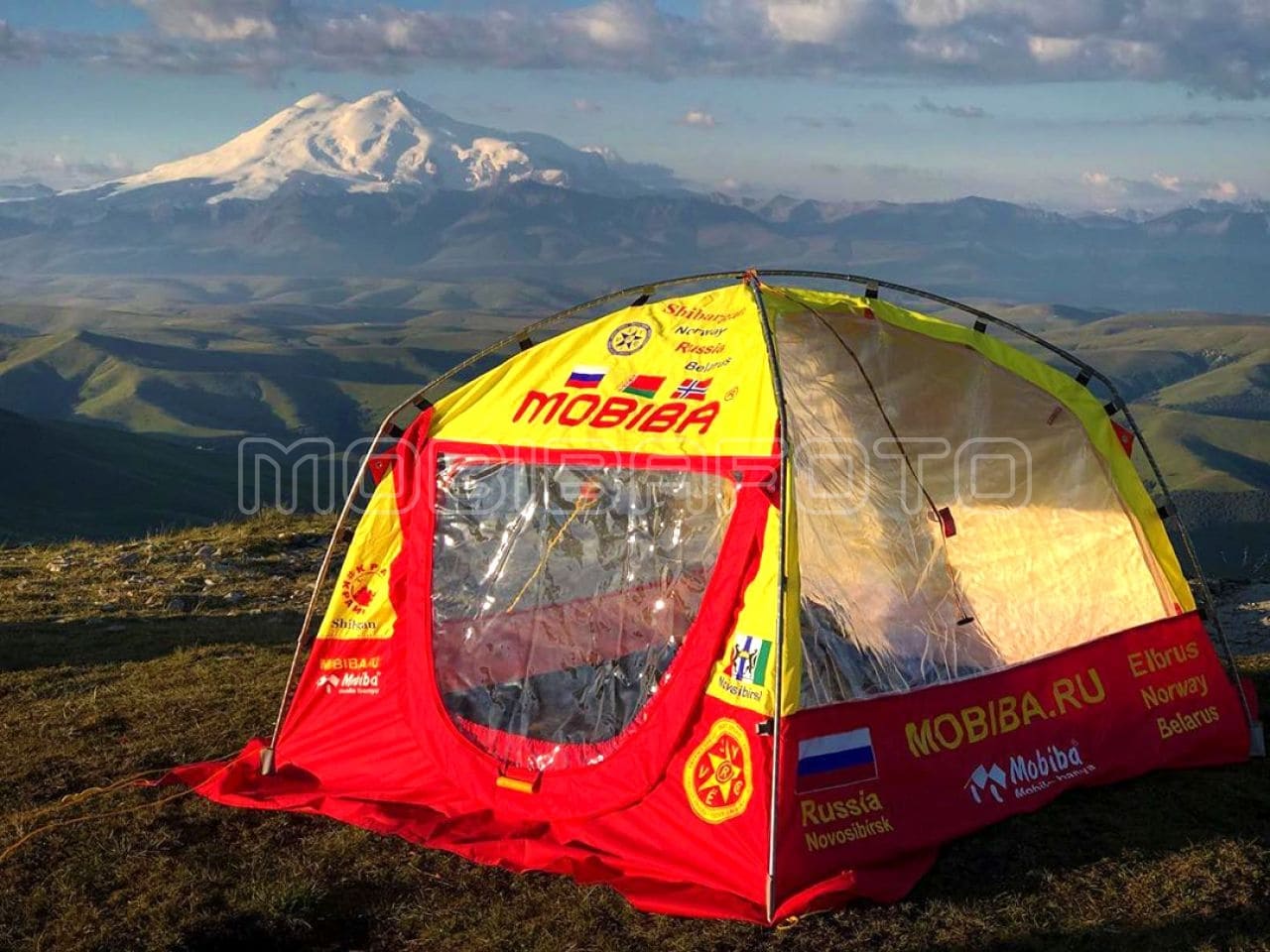 Mobiba Expeditions-Saunazelt – Elbrus-Projekt Rot-gelbes Mobiba Saunazelt mit großen transparenten Seitenfenstern und Tür auf einem Bergkamm; im Hintergrund ein verschneiter Gipfel