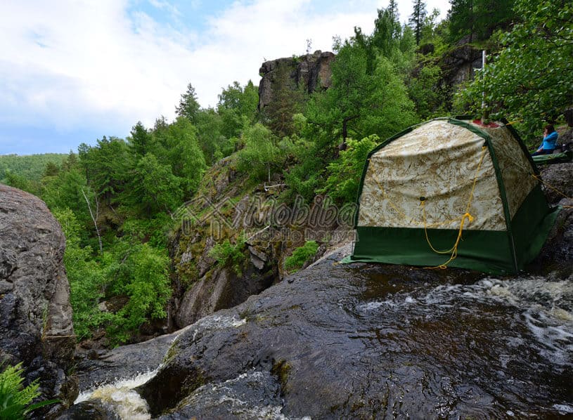 Mobiba Rucksacksauna RB-170 am Wasserfall Mobiba Rucksacksauna RB-170 auf einem Felsen neben einem Wasserfall; umgeben von Wald und felsigen Klippen.
