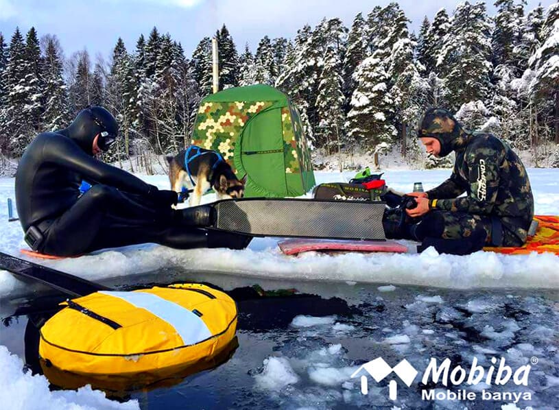 Mobiba MB-10 Zeltsauna – Aufwärmzelt für Eistaucher Zwei Taucher in Ausrüstung sitzen am Eisloch; dahinter Mobiba MB-10 Zeltsauna auf dem Eis, im Hintergrund verschneiter Wald.